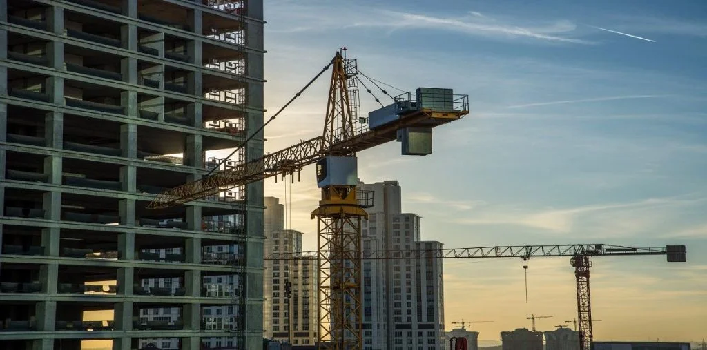 Imagem de obra em andamento, com guindaste erguendo blocos, céu azul ao fundo