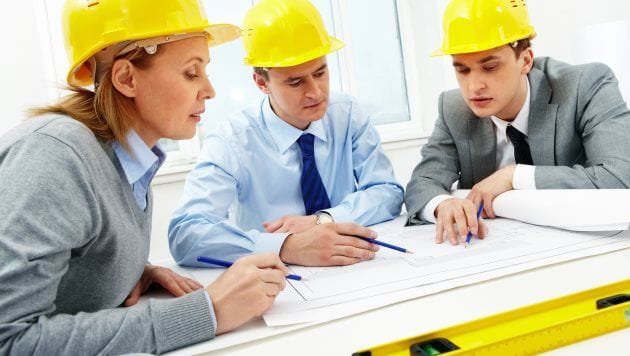 7 dicas para calouros de engenharia Três engenheiros com capacete amarelo sentados em ao redor de uma mesa analisando um projeto.