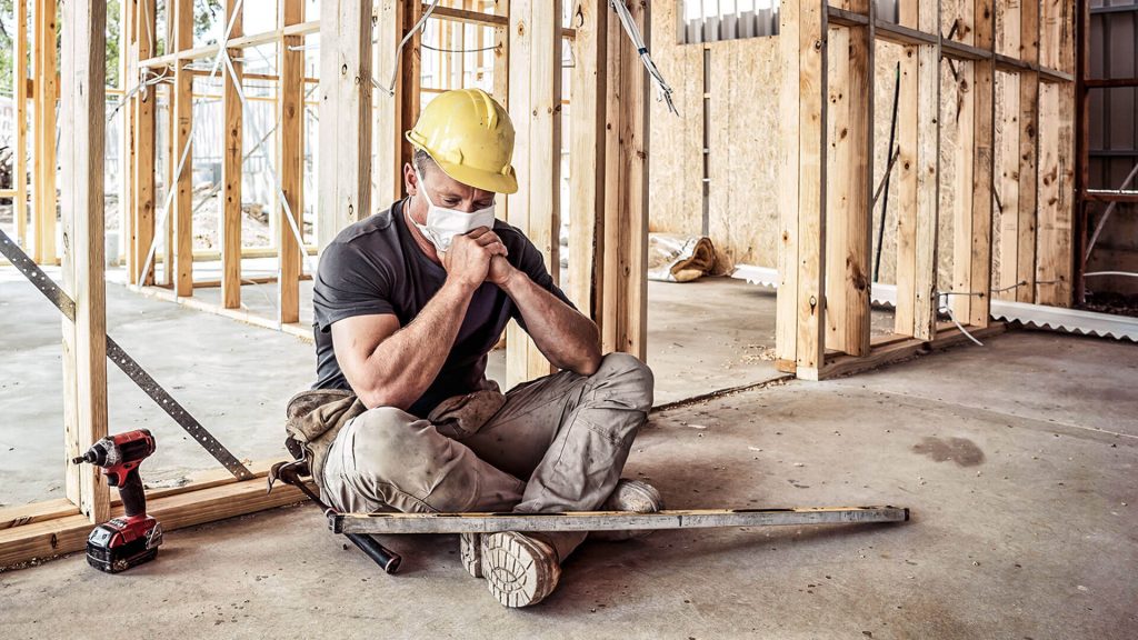 Homem com máscara e capacete em canteiro de obras sentado.