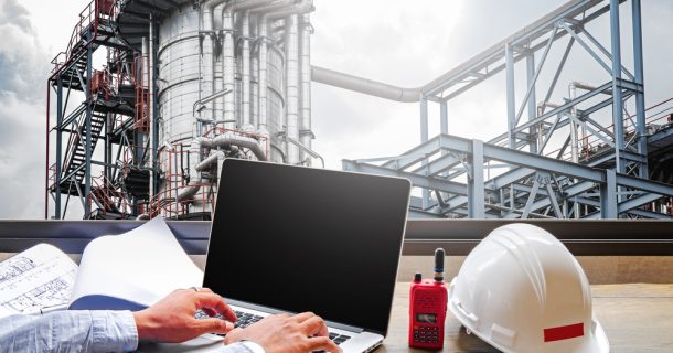 Homem utilizando um notebook ao lado de um capacete branco próximo a uma instalação industrial