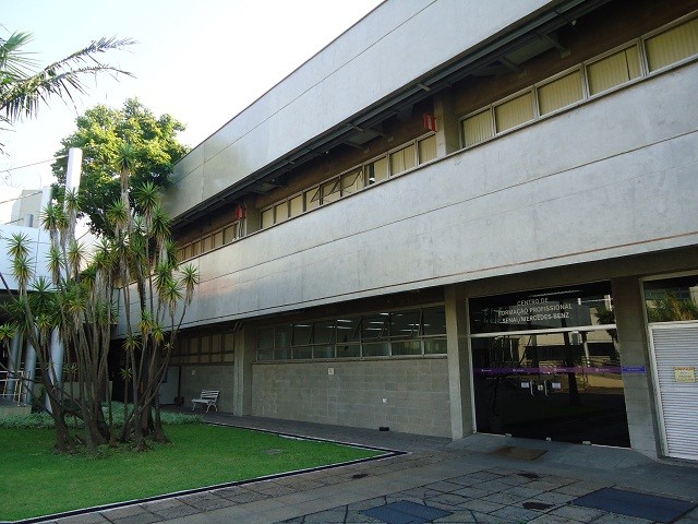 O prédio de treinamento com uma porta de vidro a direita e uma árvore a esquerda da foto