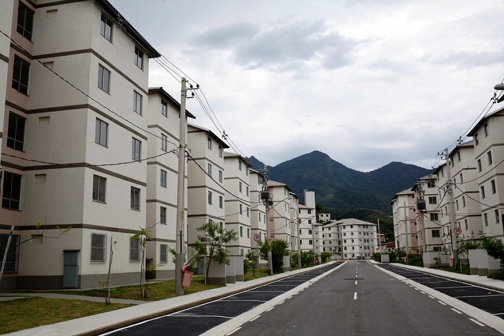 Vista de conjunto habitacional de apartamentos, com rua pavimentada entre blocos de edificações com 5 pavimentos.