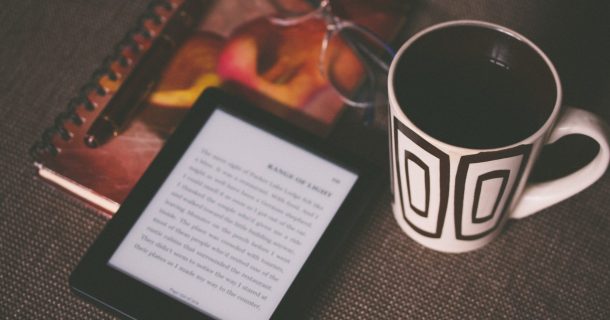 Tampo de mesa marrom escuro sobre o qual repousam um caderno com um óculos em cima, um leitor de livros eletrônico ligado com texto do ebook ilegível e uma caneca bege com desenhos abstratos em marrom escuro e café dentro.