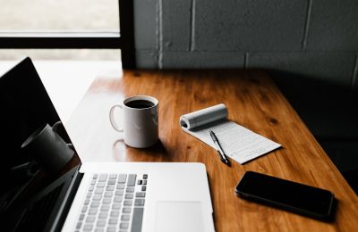 Tampo de mesa marrom sobre o qual estão um macbook pro, uma caneca branca com café, um bloco de notas e uma caneta e um celular que podem ser usados para leitura de diversos títulos.
