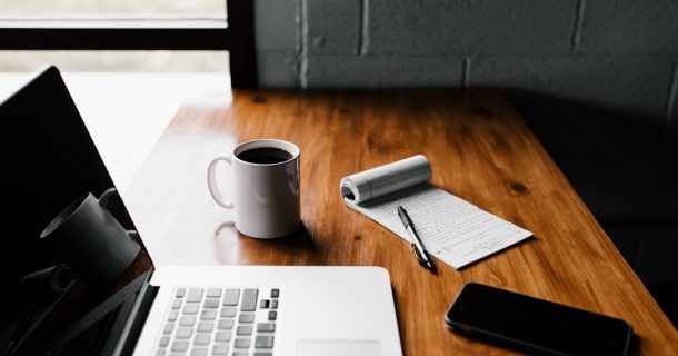Tampo de mesa marrom sobre o qual estão um macbook pro, uma caneca branca com café, um bloco de notas e uma caneta e um celular que podem ser usados para leitura de diversos títulos.