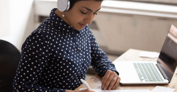 Mulher vestida com camisa de manga longa de bolinhas brancas, sentada em frente a um notebook, usando fones de ouvido externos. A imagem denota atividade de EAD.