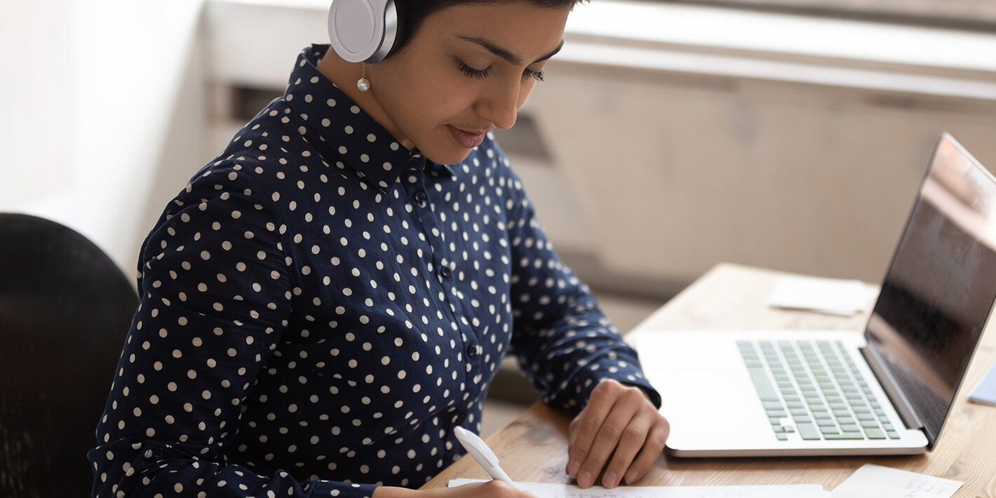 Mulher vestida com camisa de manga longa de bolinhas brancas, sentada em frente a um notebook, usando fones de ouvido externos. A imagem denota atividade de EAD.